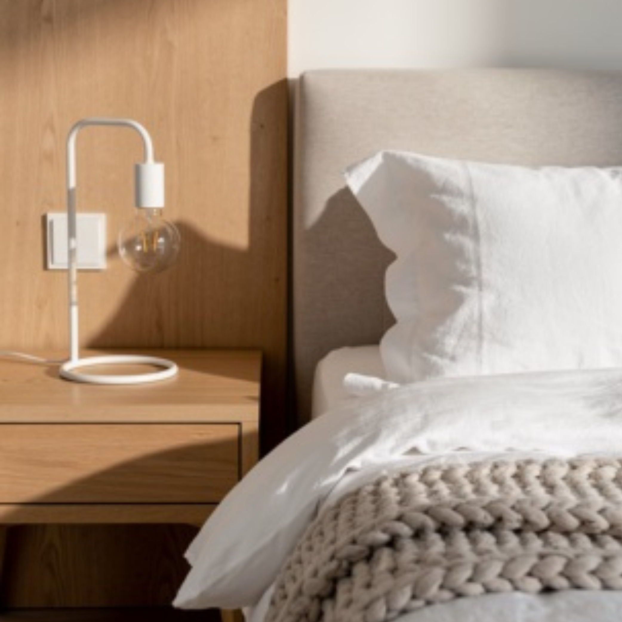 Bedroom with wooden nightstand, lamp, and bed with white pillows and beige blanket.